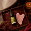 Dropper bottle of Saorse Skincare facial oil beside a pink gua sha stone on a red marble tray with a lit candle nearby.