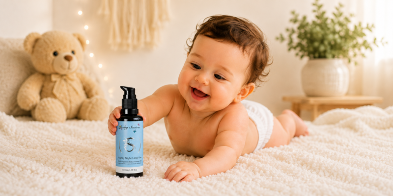 Baby crawling on a soft cream rug, reaching for a blue bottle of Baby Sage Nighty Night oil, with a teddy bear nearby.